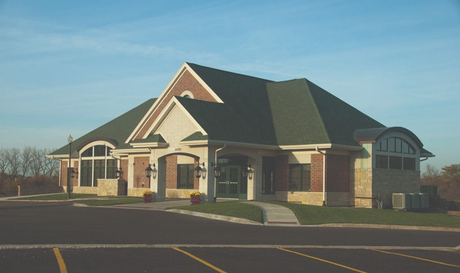 Front view of the Event Centre, a large brick building with a green roof and a main entrance. A paved sidewalk leads to the building, with grass and trees surrounding the area under a clear sky.