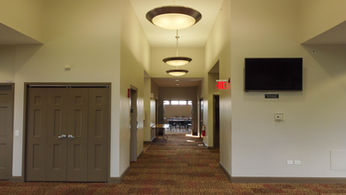 Long hallway with ceiling lights, EXIT sign, doors, and TV screen.