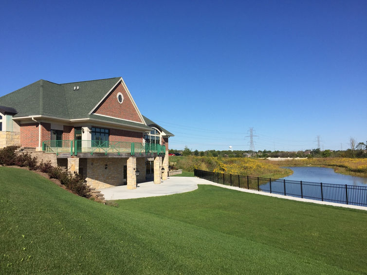 A two-story brick building called the Event Centre with a green roof that sits on a grassy hill next to a small pond. A sidewalk and black metal fence run along the water in front of the building under a clear blue sky.