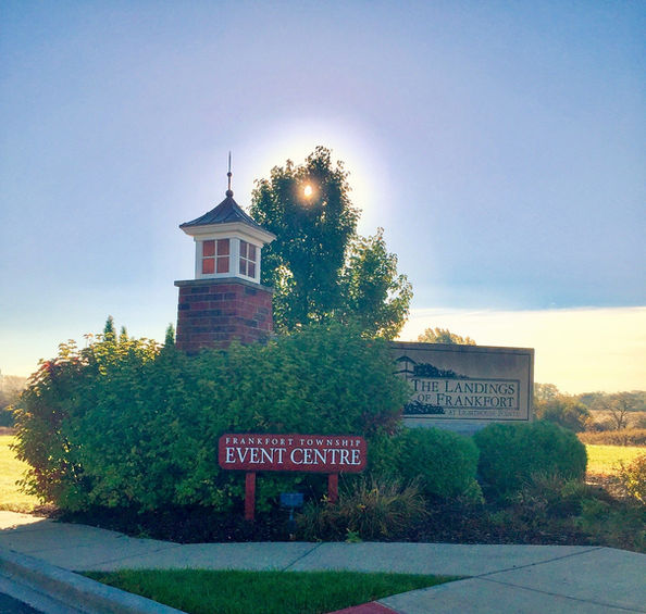 Entrance sign for the Frankfort Township Event Centre surrounded by green shrubs and landscaping, with a brick pillar topped by a small lighthouse-style structure. Behind the bushes is another sign reading “The Landings of Frankfort,” with sunlight shining through a tree and a sidewalk in the foreground.